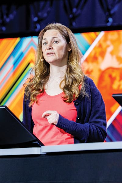 A woman is speaking at a conference. She stands behind a desk with monitors, dressed in a coral shirt and jeans. The backdrop is a colorful, geometric design with a lit stage above.