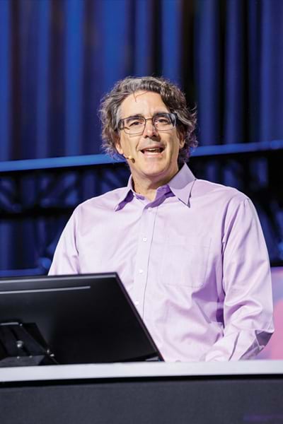 A man speaking at a conference. He has curly gray hair, glasses, and is wearing a purple shirt.