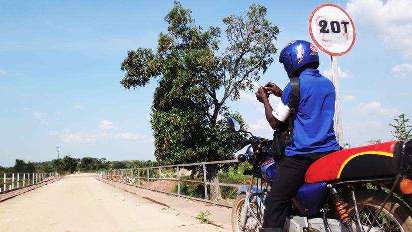 A man wearing a blue shirt and helmet sits on a motorcycle on a bridge in Africa. He's looking at a small device in his hands. A sign reads "20T." Trees and a blue sky with light clouds are in the background.