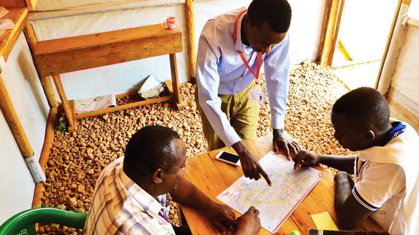 Three people looking at a map on a table in a tent-like structure. One person stands and points to the map. Another sits to the left and a third sits to the right. Scissors and other objects are on the table.