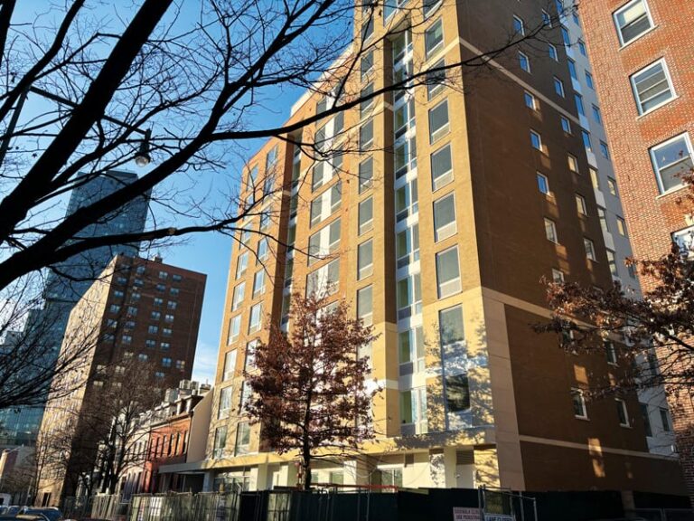 A street scene with several tall brick buildings under a clear sky, with a 10-story building in the foreground. Bare tree branches frame the buildings. Construction fencing is at the base of the closest building.