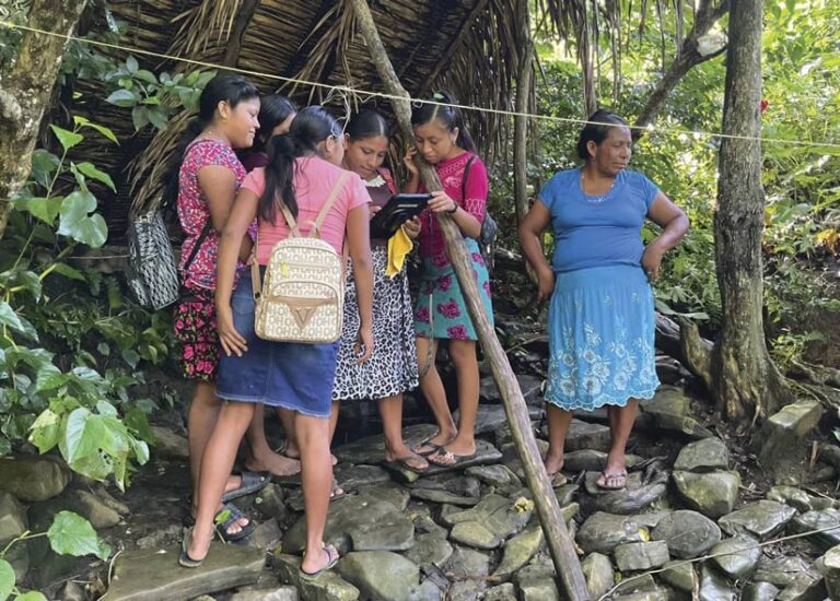 Six women stand in a forested area. One is looking at a tablet. They are standing on rocks that have water running over them.