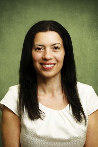 A woman with long black hair and a bright smile wearing a white patterned shirt against a green background.