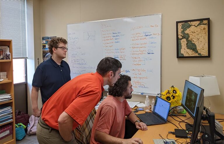Three people look at computer screens in an office. A whiteboard behind them is full of notes and drawings. A framed map of the San Francisco Bay Area hangs on the wall.