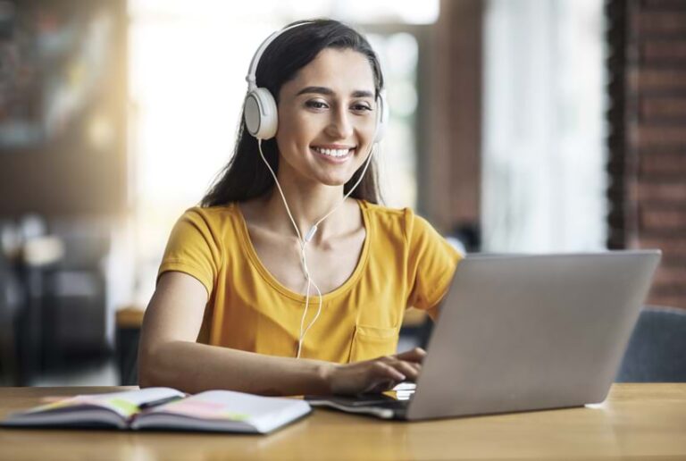 A young woman in a yellow shirt works on a laptop at a table in a room with a window.