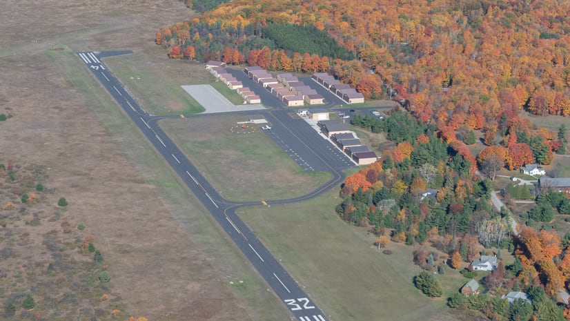 An aerial view of an airport runway marked 14/32. The runway is surrounded by grass, with a row of buildings and fall foliage on one side and sparse vegetation on the other.