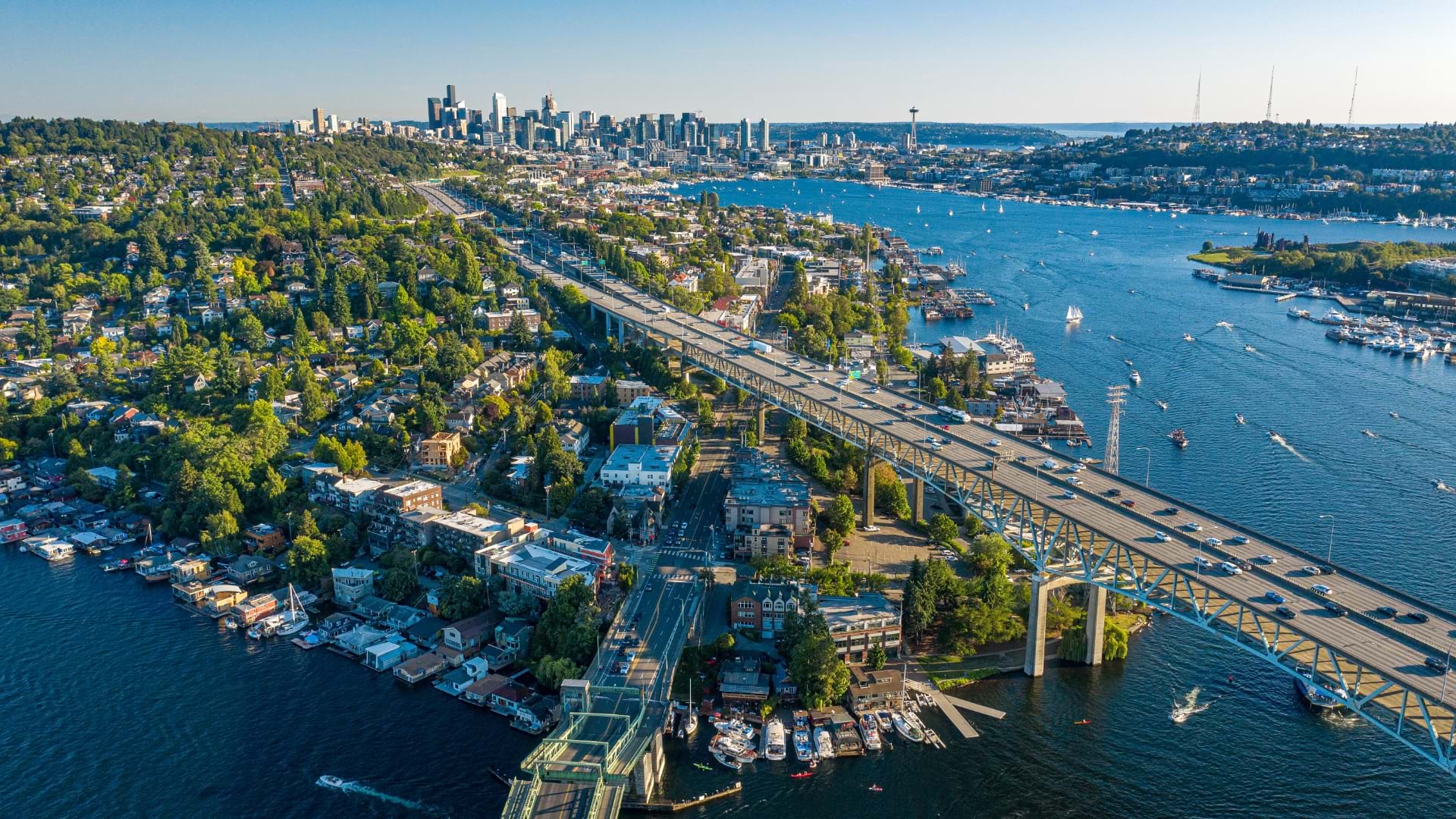 Aerial view of Seattle showing the Aurora Bridge spanning the Lake Union ship canal, with downtown Seattle's skyline in the background and residential neighborhoods on tree-covered hills. The image captures Seattle's constrained geography with water bodies on multiple sides, limited bridge crossings, and dense urban development that creates natural traffic bottlenecks during major events.