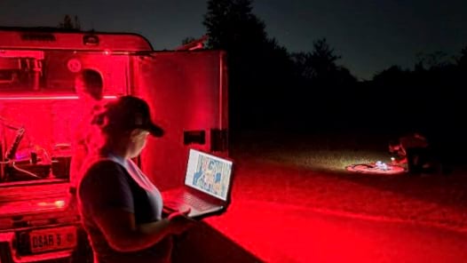 Nighttime scene showing search and rescue team member using laptop computer with thermal imaging interface next to mobile command vehicle under red tactical lighting, with thermal-equipped drone operations visible in background