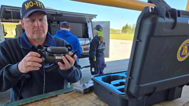 Chad Sperry wearing MO-TF1 baseball cap operating drone controller equipment at outdoor training site with team members and mobile command vehicle visible in background