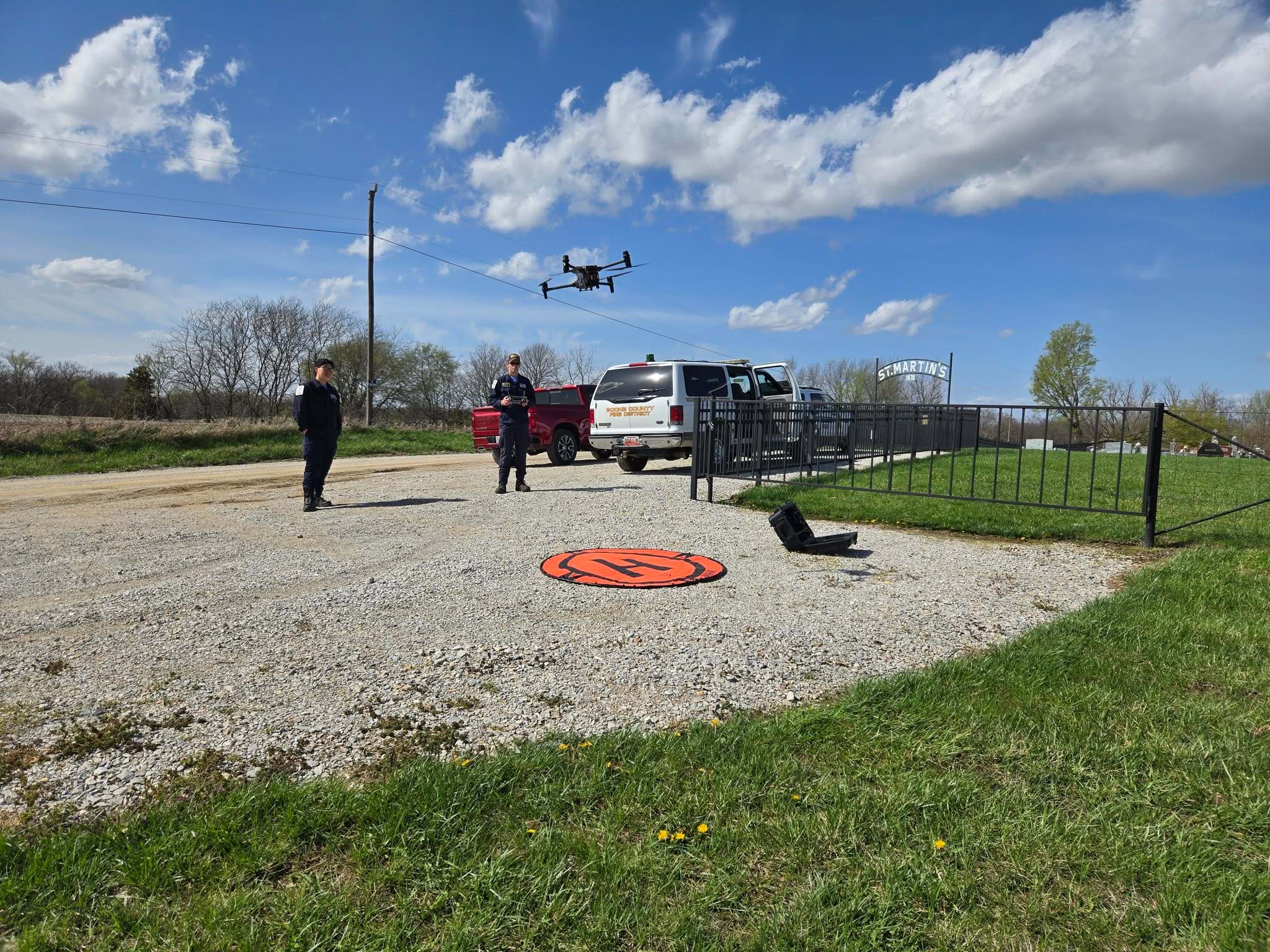 Drone hovering above orange circular landing pad in grassy field with two uniformed operators nearby, emergency vehicles in background, and safety fencing visible under partly cloudy sky