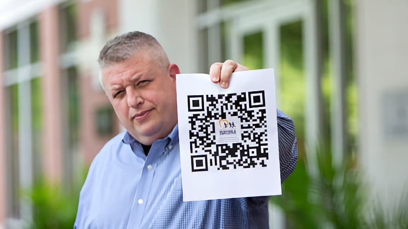 John E. Bischoff III holds up a white paper with a black QR code, standing outside a modern building with large windows and greenery in the background.