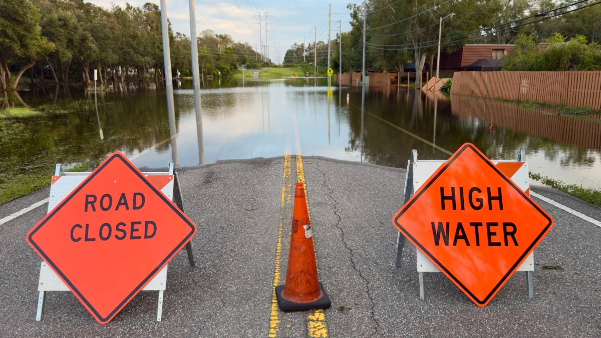 Flooded residential street with "Road Closed" and "High Water" warning signs, showing water covering road and surrounding properties.