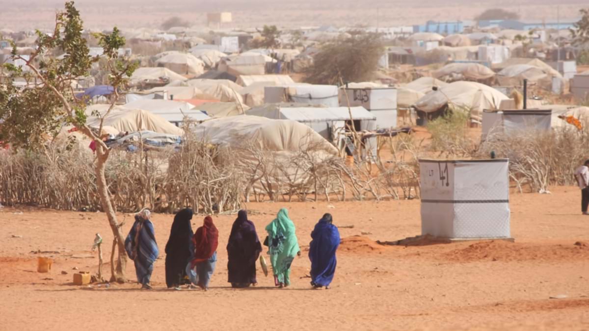 Five women in colorful traditional clothing walk across arid ground past rows of beige and white tents and makeshift shelters in a displacement camp in Mali, with sparse vegetation in the foreground.
