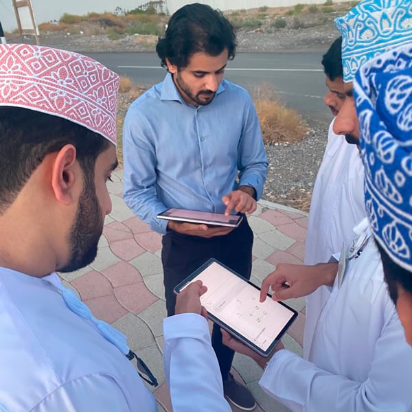 Photo of several men on a sidewalk gathered around two tablets, apparently conversing with one another.