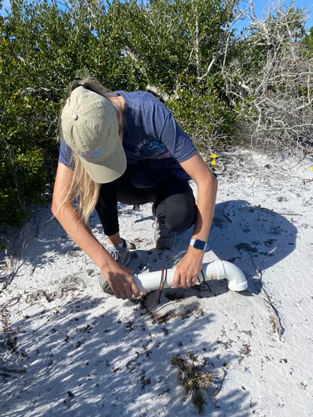 A photo of a woman wearing a baseball cap with a short length of white PVC piping in her hands.