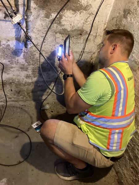 Photo of a man in a high-visibility vest crouched in what appears to be an unfinished basement. The man is holding a tablet and a light up to a pipe coming out of a wall.