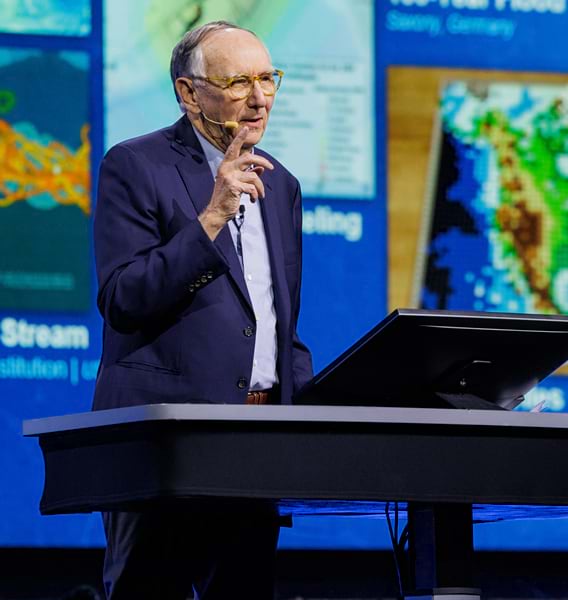 Photo of Jack Dangermond speaking with a presentation on a large screen behind him, with one finger raised in the air.