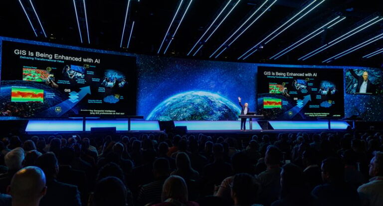 Photo of Jack Dangermond speaking with a presentation on a large screen behind him, with one hand raised and a huge crowd of people seated in the foreground.