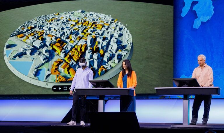 Three presenters stand at monitors in front of a huge display. One of the presenters is wearing a VR headset. The display shows a 3D circular cutout of a city, with many buildings colored orange.