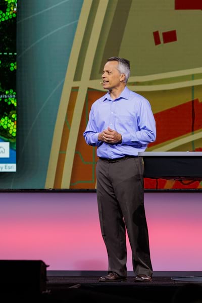 Photo of a man standing onstage, speaking with his hands clasped.