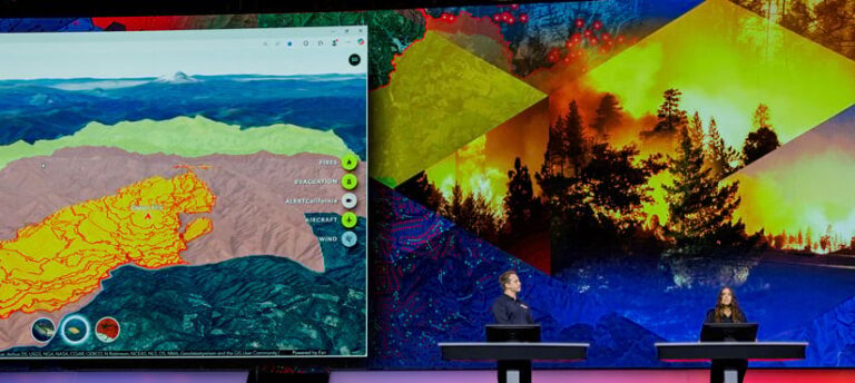 Two presenters stand at monitors onstage with a graphic of a map representing wildfires on the display behind them.