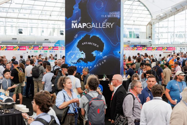 Photo of a huge crowd of people talking to each other at an event, with a large blue and black map display set up in the middle of the floor. The display reads “Esri Map Gallery.”