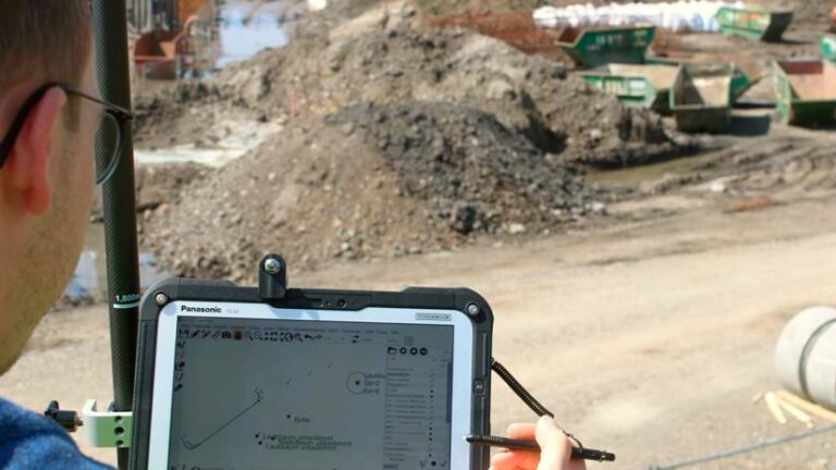 Photo of a man with his back to the camera, holding a tablet so the screen is visible, with debris and construction materials are in the background.