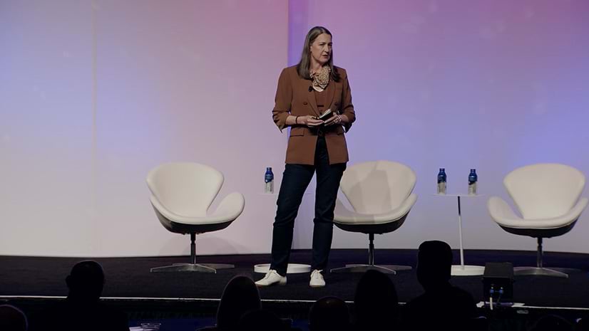 A person wearing business casual attire standing on a presentation stage backlit with a light purple gradient