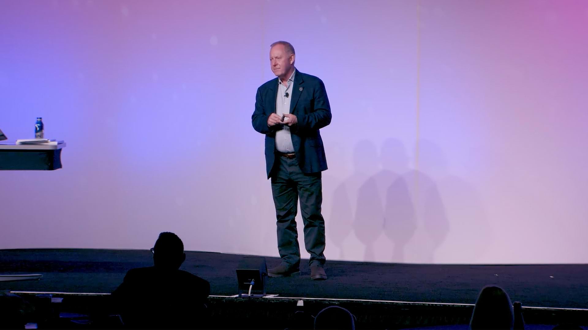 A person wearing a business suit standing on a presentation stage backlit with a light purple gradient