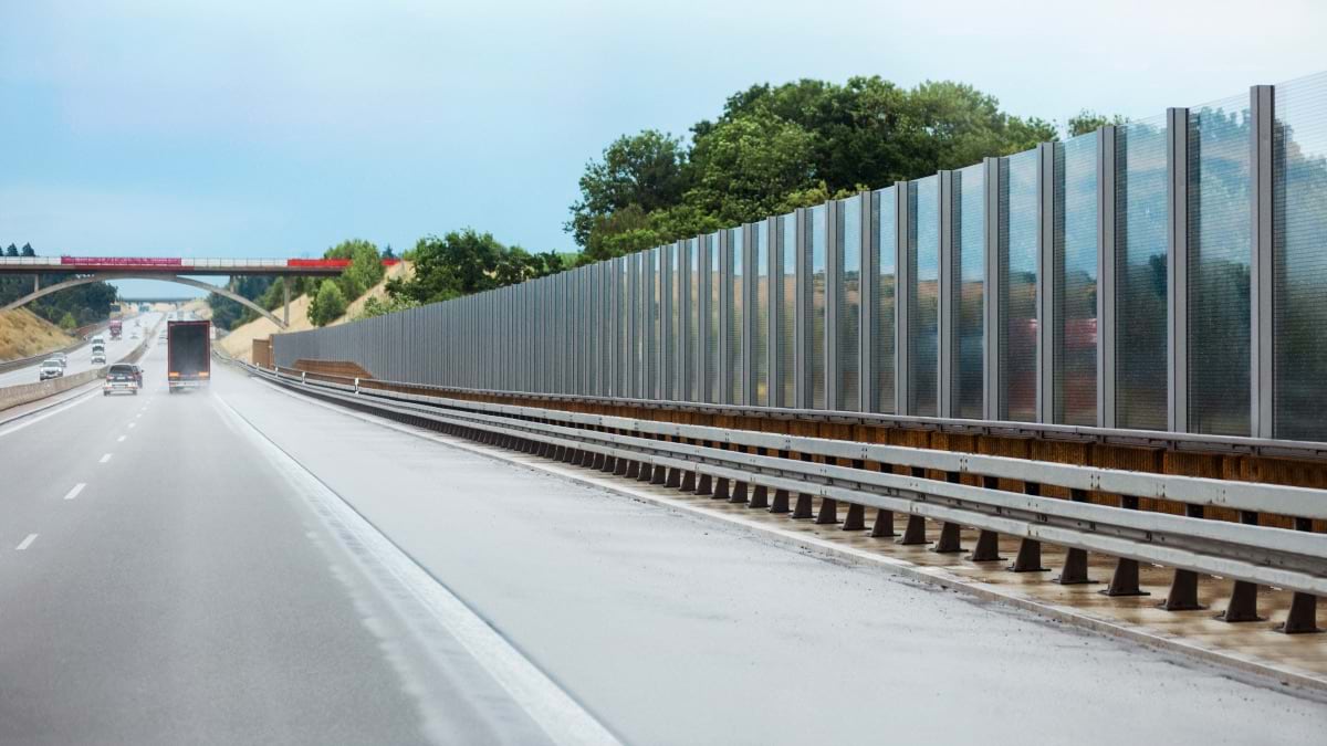 Tall transparent noise barrier wall along European highway with cars and residential buildings visible beyond, showing noise pollution mitigation