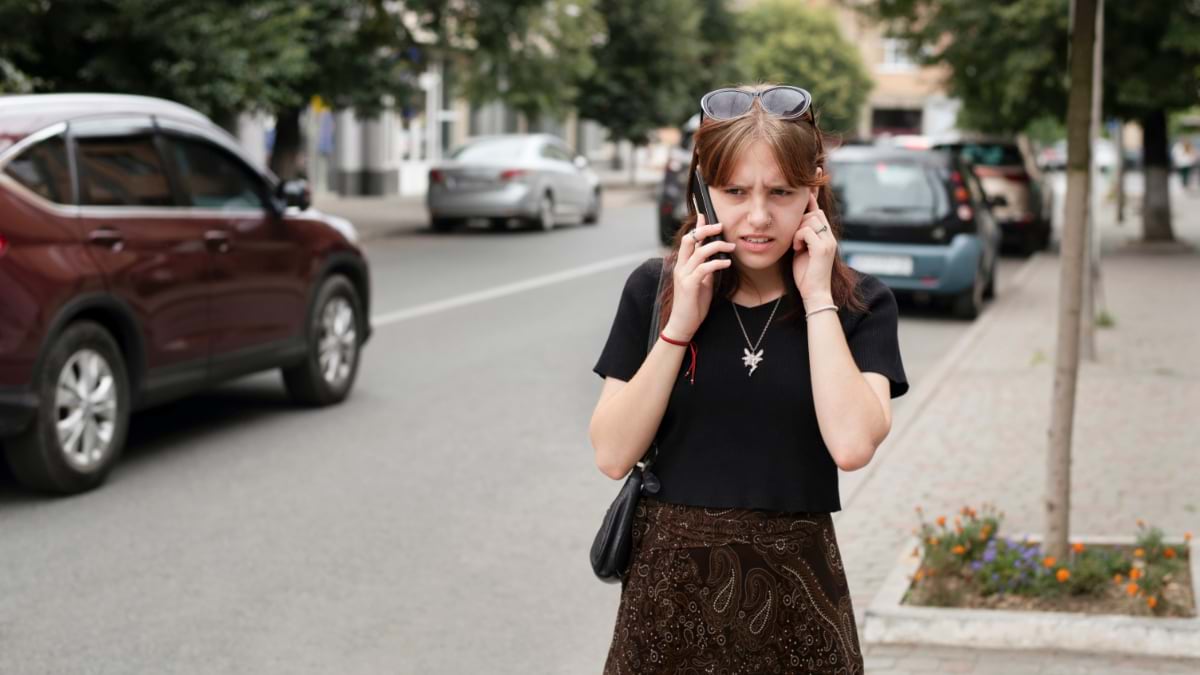 Woman on phone covering ear from traffic noise on busy street with cars in background, demonstrating urban noise pollution exposure in Europe