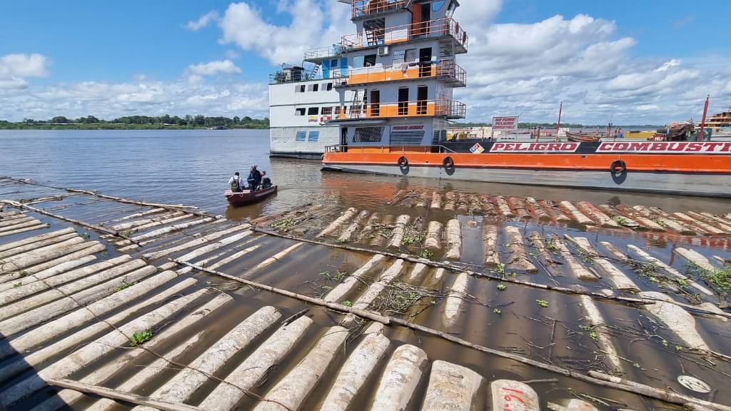 Rows of logs organized on a barge beside muddy river water, with police patrol boats and officers visible in background.