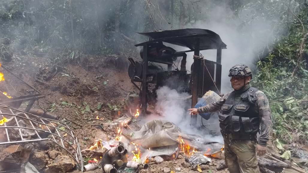 Uniformed tactical officer standing beside burning mining equipment with flames and smoke in jungle clearing.