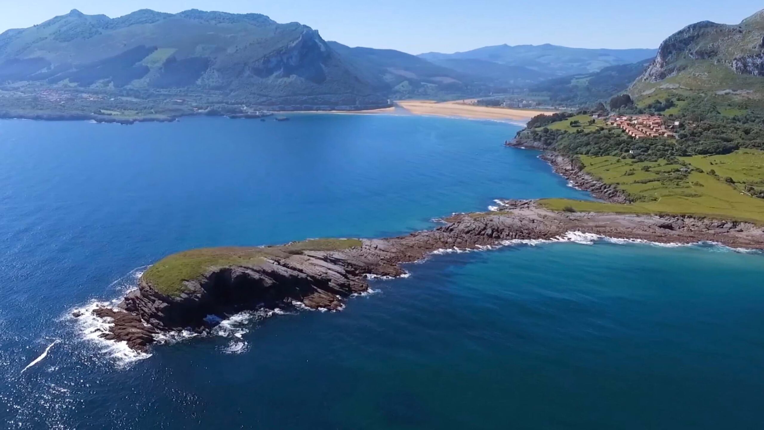 Aerial view of Cantabria's rocky coastal peninsula with turquoise waters, sandy beaches, and mountain ranges in the background