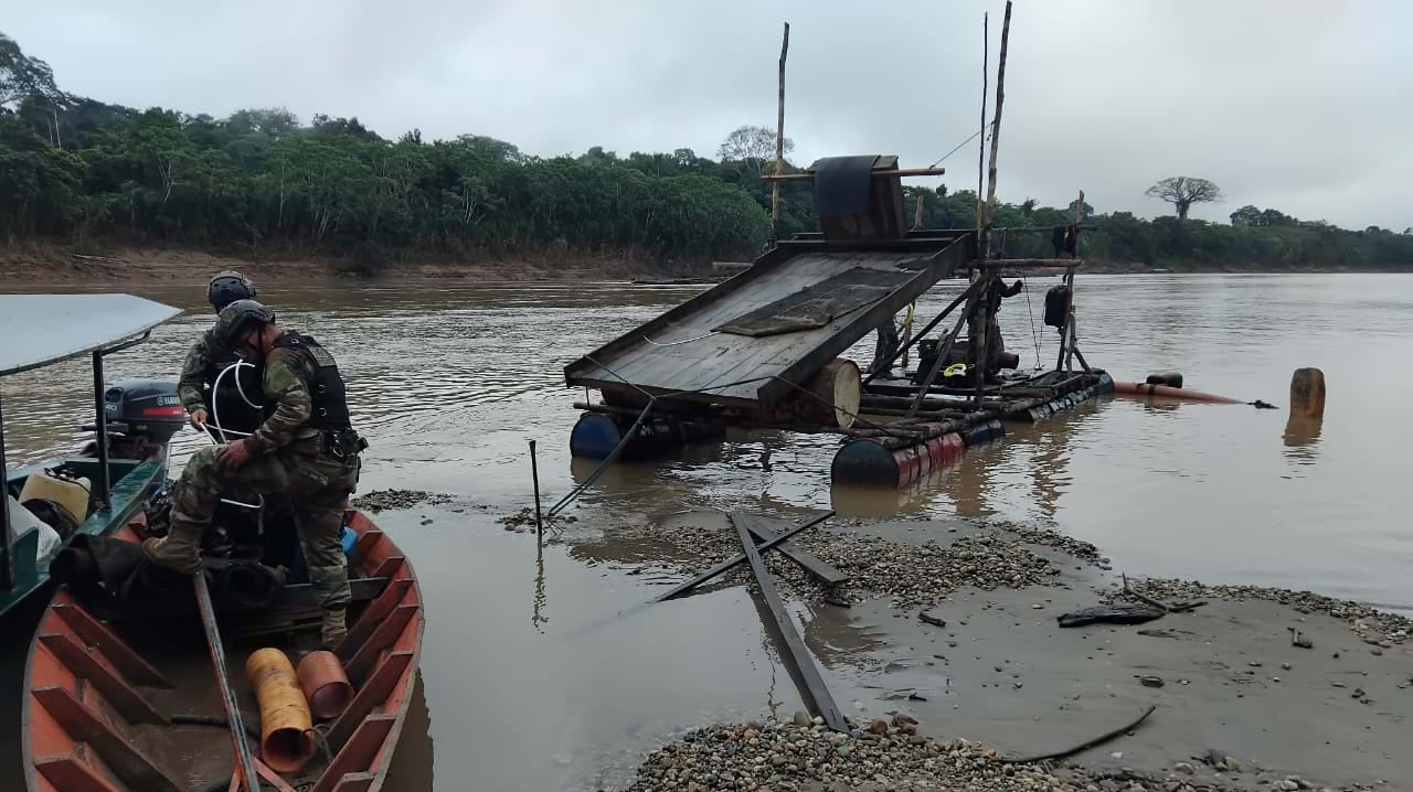Military personnel in patrol boat observing partially submerged illegal mining dredge with blue flotation barrels in muddy Amazon river, dense rainforest visible on horizon.