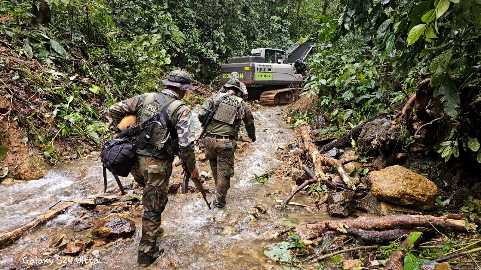 Two officers in camouflage uniforms approaching yellow CAT excavator concealed among dense tropical vegetation and fallen logs.
