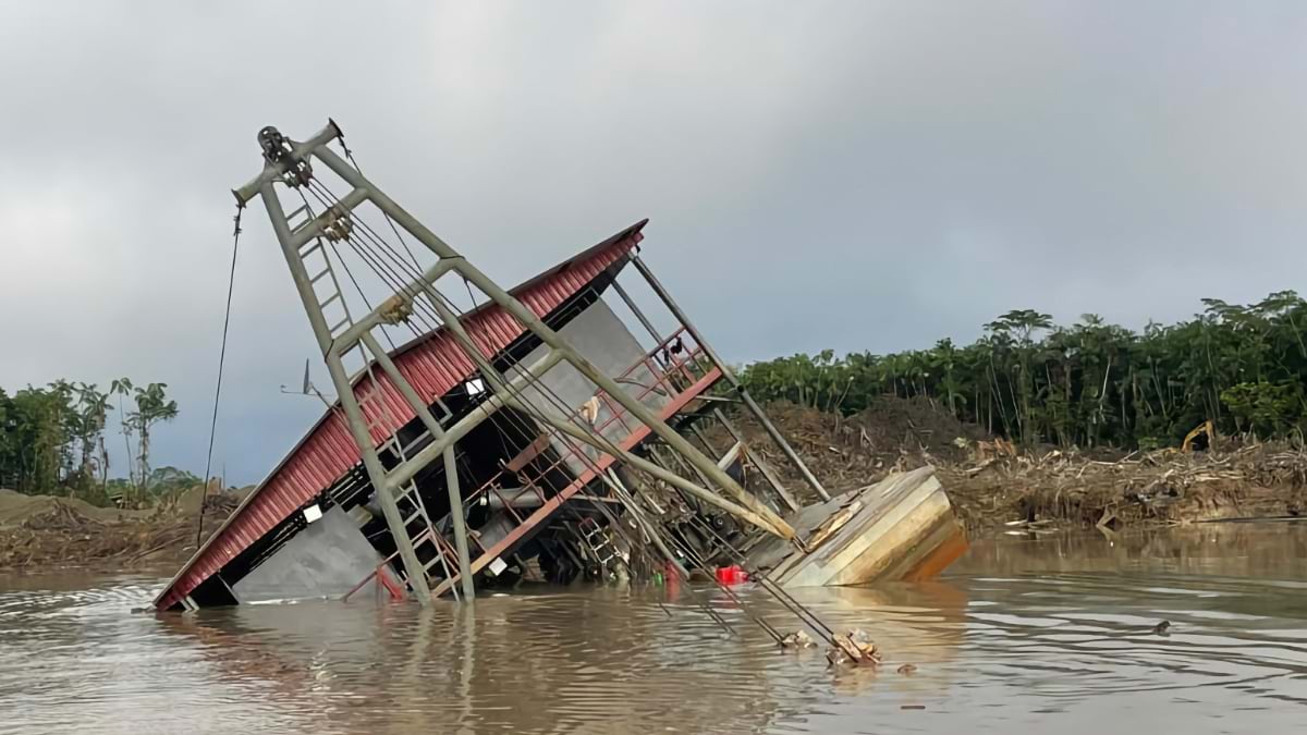 Large industrial mining vessel tilted and sinking in muddy brown river water with deforested landscape visible in background.