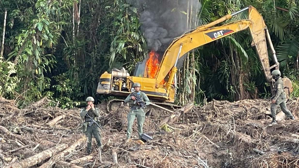 Yellow construction excavator fully engulfed in fire with armed military officers standing guard in cleared forest area surrounded by smoke and debris.