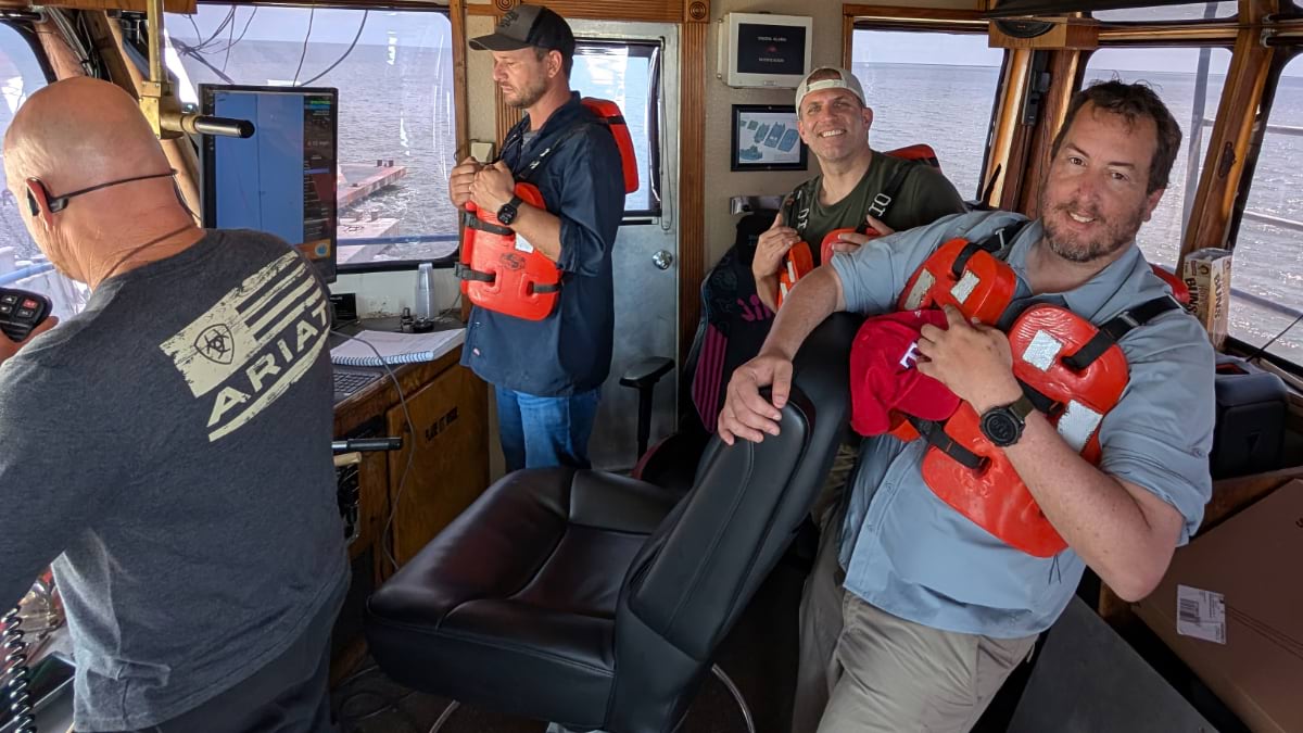 our crew members in orange life vests gather in boat wheelhouse with navigation screens visible, blending traditional fishing work with digital mapping.