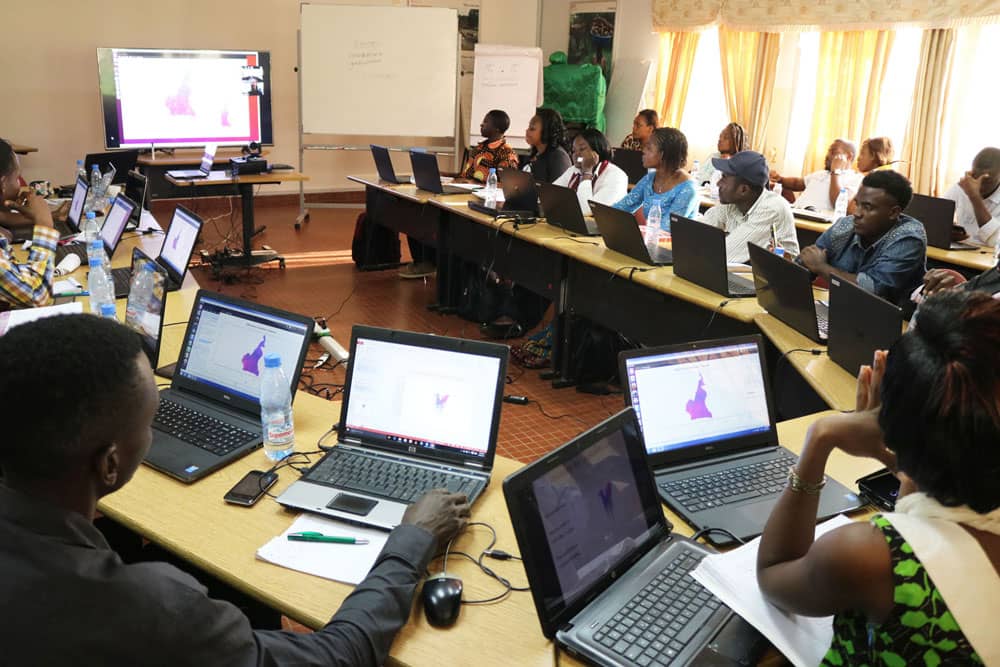 Workshop participants at laptops displaying purple-toned geographical maps during Congo Basin Institute GIS training session in Cameroon classroom.