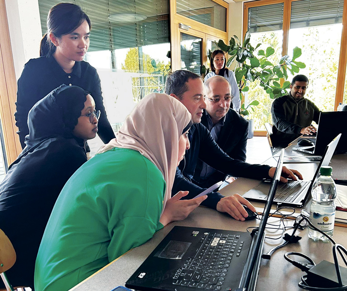A diverse group of professionals gather around a table, working together on laptops in a bright, modern office.