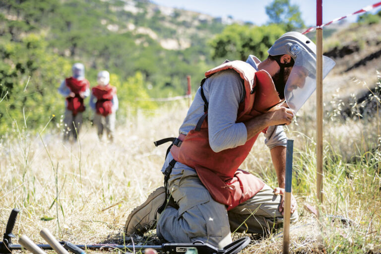 A demining expert in protective gear examines the ground, with two more experts blurred in the background.
