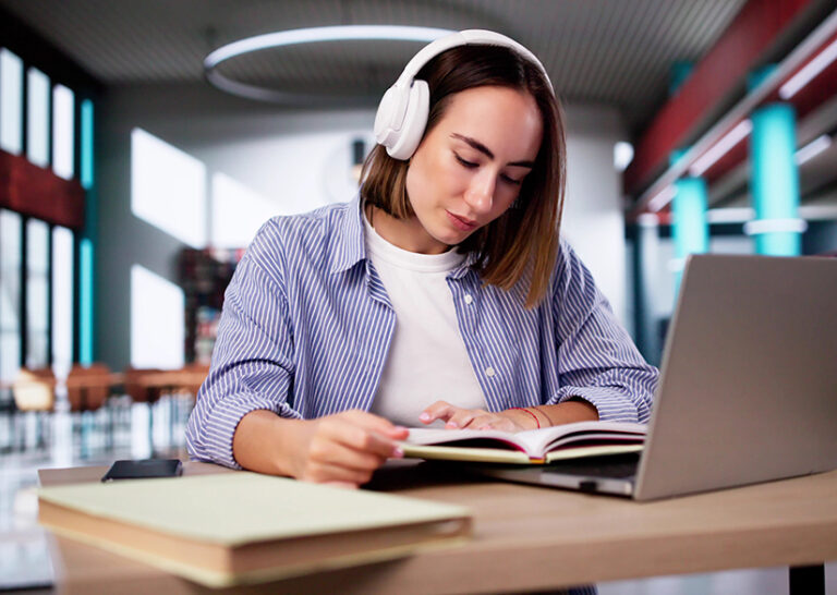 A woman with headphones studies in a library with a book and a laptop.