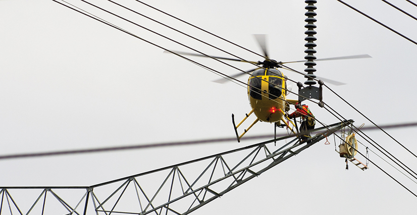 A yellow helicopter hovers near power lines while a worker performs maintenance on the tower.