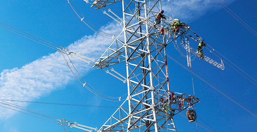 Workers repair a high-voltage transmission tower against a partly cloudy blue sky. The tower's metal lattice and wires create a complex geometric pattern.