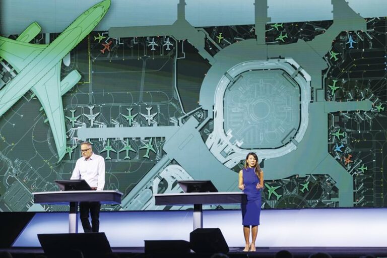 A man and woman stand behind podiums giving a presentation on a stage. Behind them is a large screen depicting an airport diagram with airplanes taxiing and parked. The woman is wearing a blue dress, and the man is wearing a white shirt.