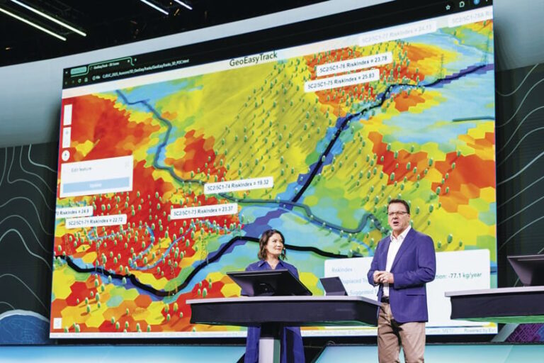 A wide shot of a presentation stage with a man in a blue blazer speaking near a woman at a podium. A large screen behind them displays a map with hexagonal sections colored from red to blue. Lines representing utility infrastructure cut through the map. Small tree icons are distributed throughout.