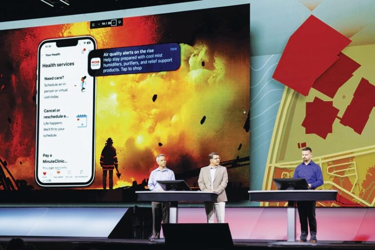Three speakers on a stage stand at podiums, in front of a large screen. The left side of the screen shows a mobile health app with the message "Air quality alerts on the rise." The app shows features to schedule appointments, manage appointments, and pay for MinuteClinic. The background shows a firefighter standing in front of large fire. A map is on the right side of the screen.