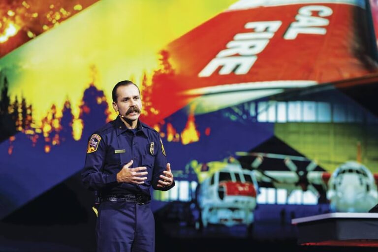A mustachioed man in a navy-blue CAL FIRE uniform stands speaking on stage. Behind him, a large screen displays an image of a burning forest. The tail of a CAL FIRE airplane is visible, with "CAL FIRE" in white letters. In the background, there are two out-of-focus helicopters.
