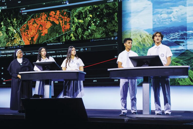 A wide shot shows five people standing behind presentation podiums. An older woman in a nun's habit stands next to two students in school uniforms. Another pair of students in uniforms are on the right. The projection screen behind them shows satellite images and graphics.